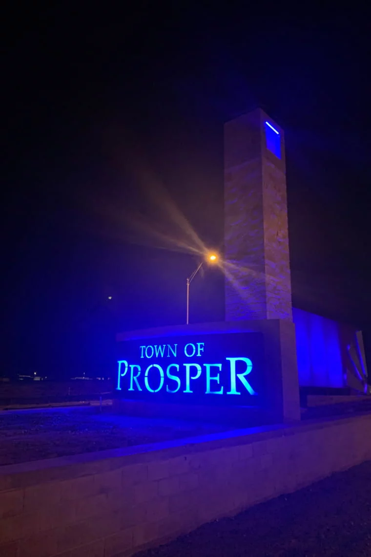 Blue illuminated sign reading 'Town of Prosper' at night next to a stone tower and streetlight.
