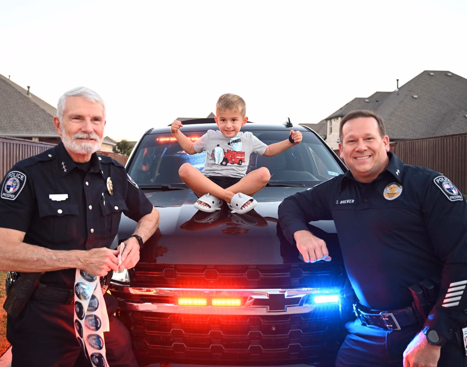 Two police officers flank a smiling boy sitting on a police vehicle with flashing red and blue lights.