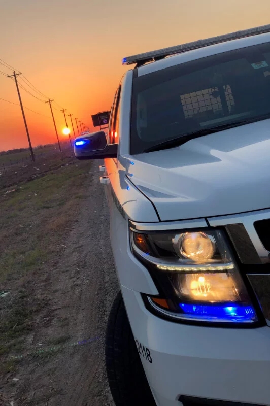 Front side view of a white police vehicle with blue emergency lights on at sunset on a rural road.
