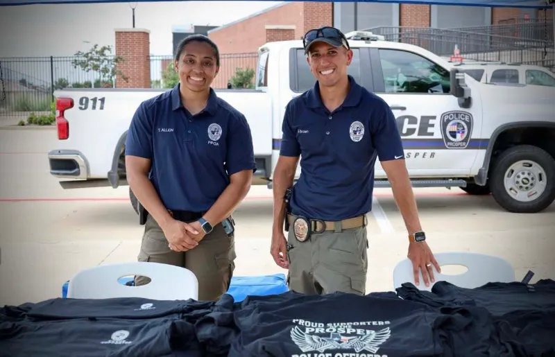 Three police officers standing in front of a Prosper Police Department tent with people at a table nearby.