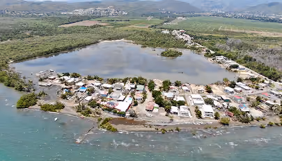 Aerial view of a coastal village by a lagoon surrounded by greenery and hills in the distance.