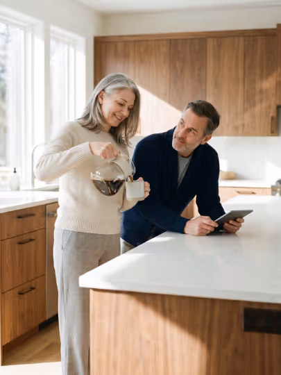 Woman pouring coffee into a cup while a man looks on holding a tablet in a bright kitchen.
