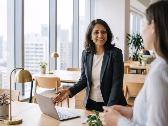 Woman in a blazer smiling and gesturing at a laptop during a meeting in a bright office.