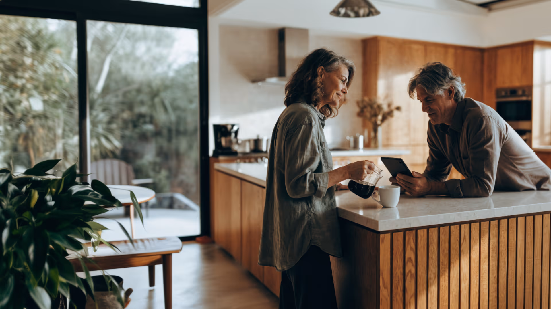 Couple having coffee in a kitchen, one pouring coffee while the other looks at a tablet.
