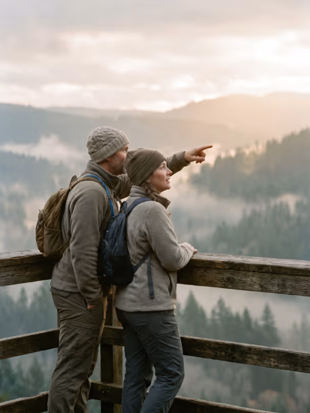 Couple wearing backpacks and hats on a wooden balcony overlooking misty forested mountains at sunrise.