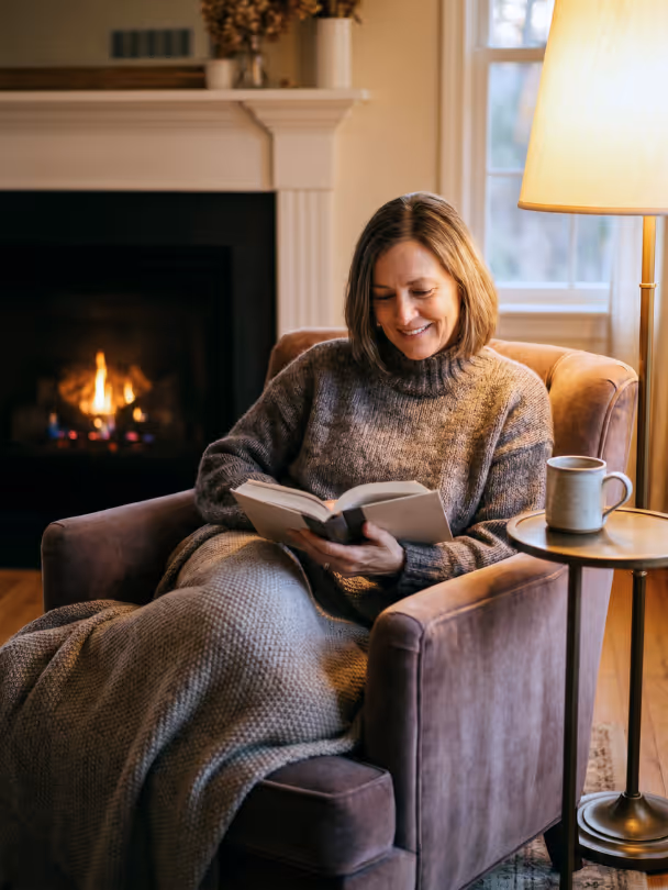 Woman in a sweater reading a book in a cozy armchair near a glowing fireplace and lamp.