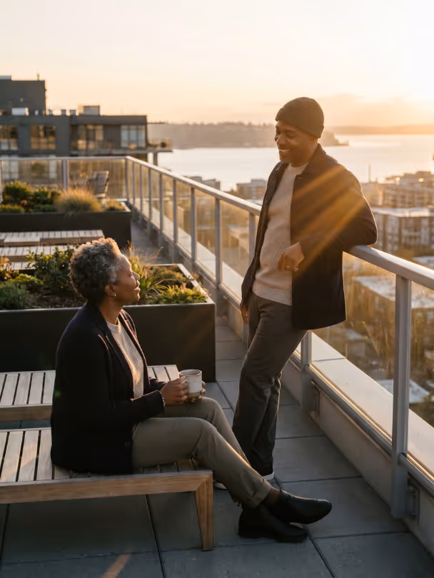 Couple enjoying sunset conversation on rooftop balcony with city and water views.
