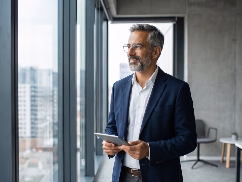 Middle-aged man in glasses and suit holding a tablet, looking out office window smiling.