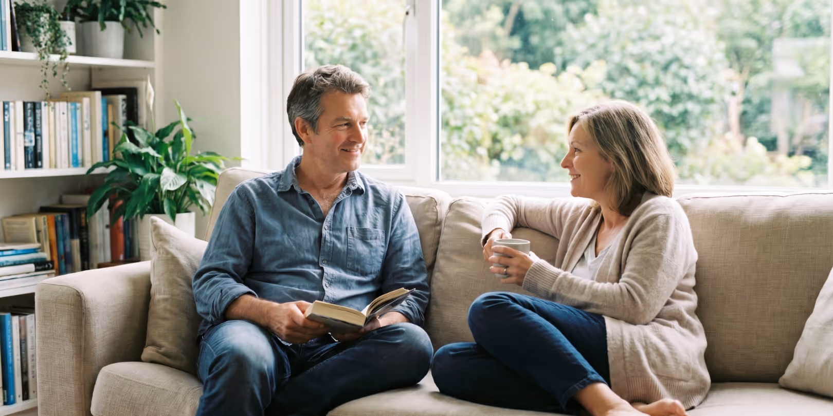 Man reading a book and woman holding a cup, sitting and smiling on a beige couch by a window.