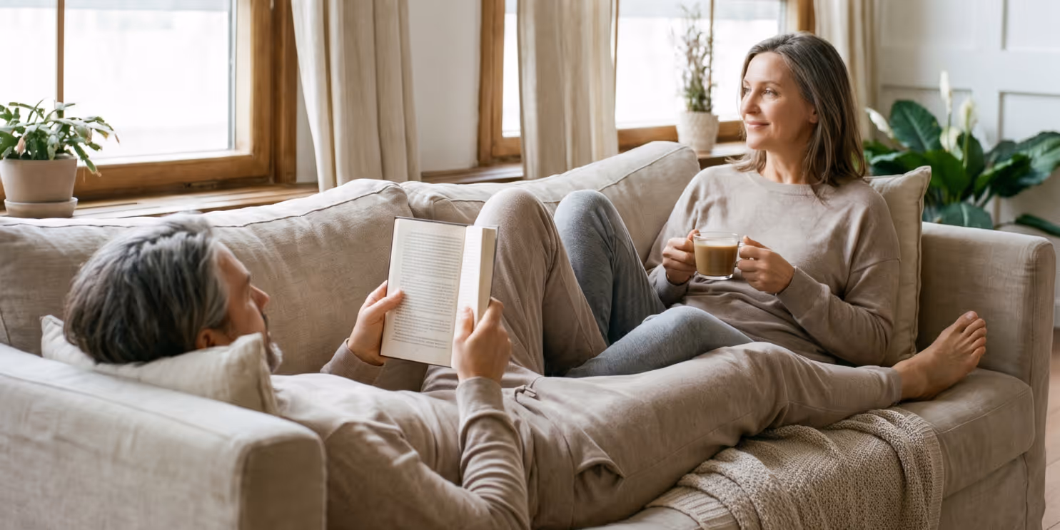 Middle-aged couple relaxing on beige couch; man reads a book while woman holds a cup, smiling.