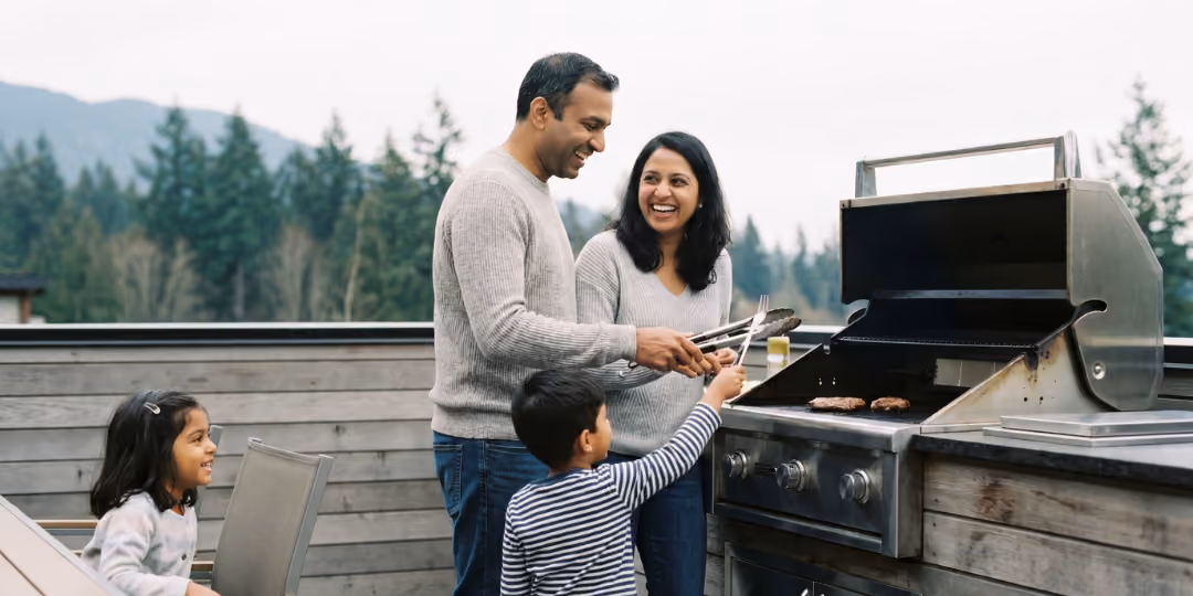 Family of four outside grilling burgers on a patio grill surrounded by trees and mountains.