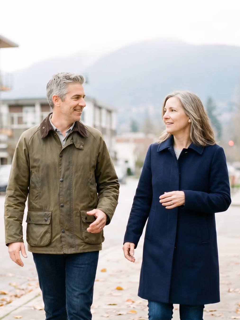 Man and woman wearing coats walking and smiling at each other on a city sidewalk.