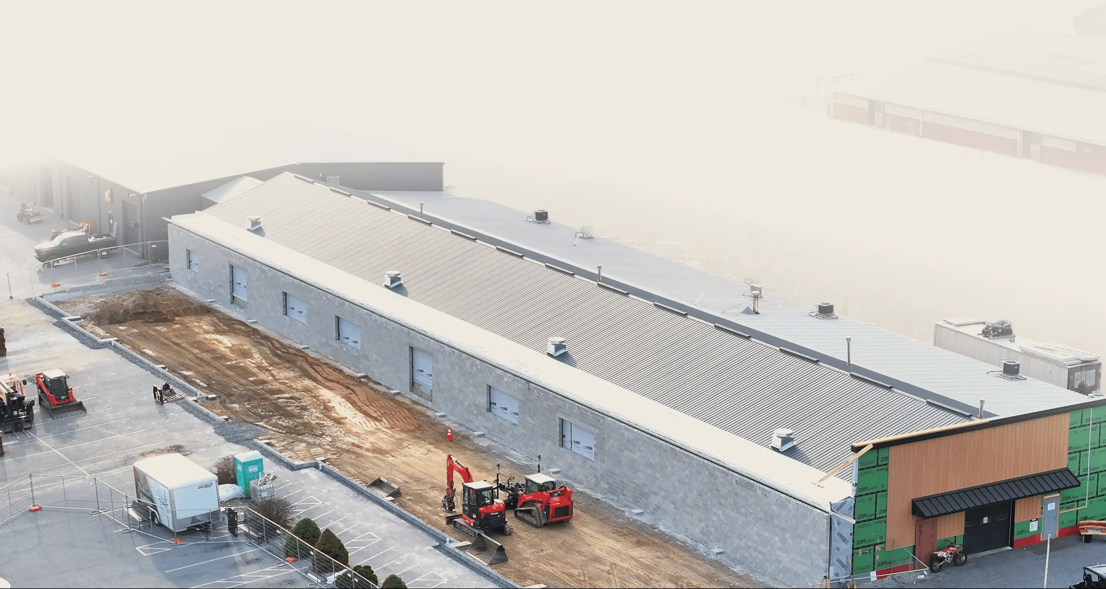 A large industrial building under construction with construction vehicles and equipment on-site, surrounded by parking areas and partially completed landscaping.