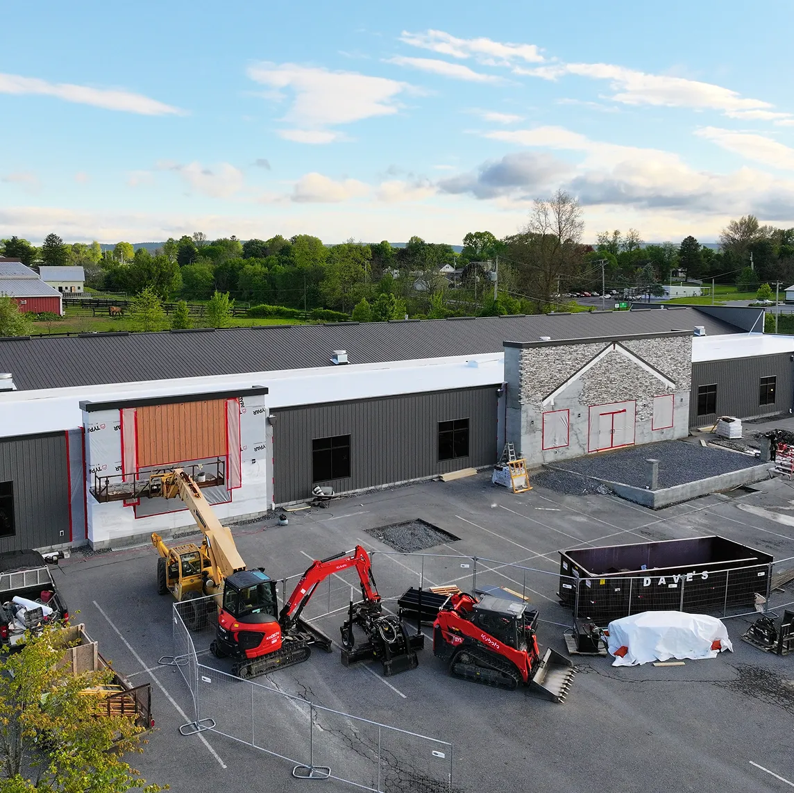A construction site with machinery, including an excavator and skid steer, in front of a partially completed commercial building with black and white exterior walls.