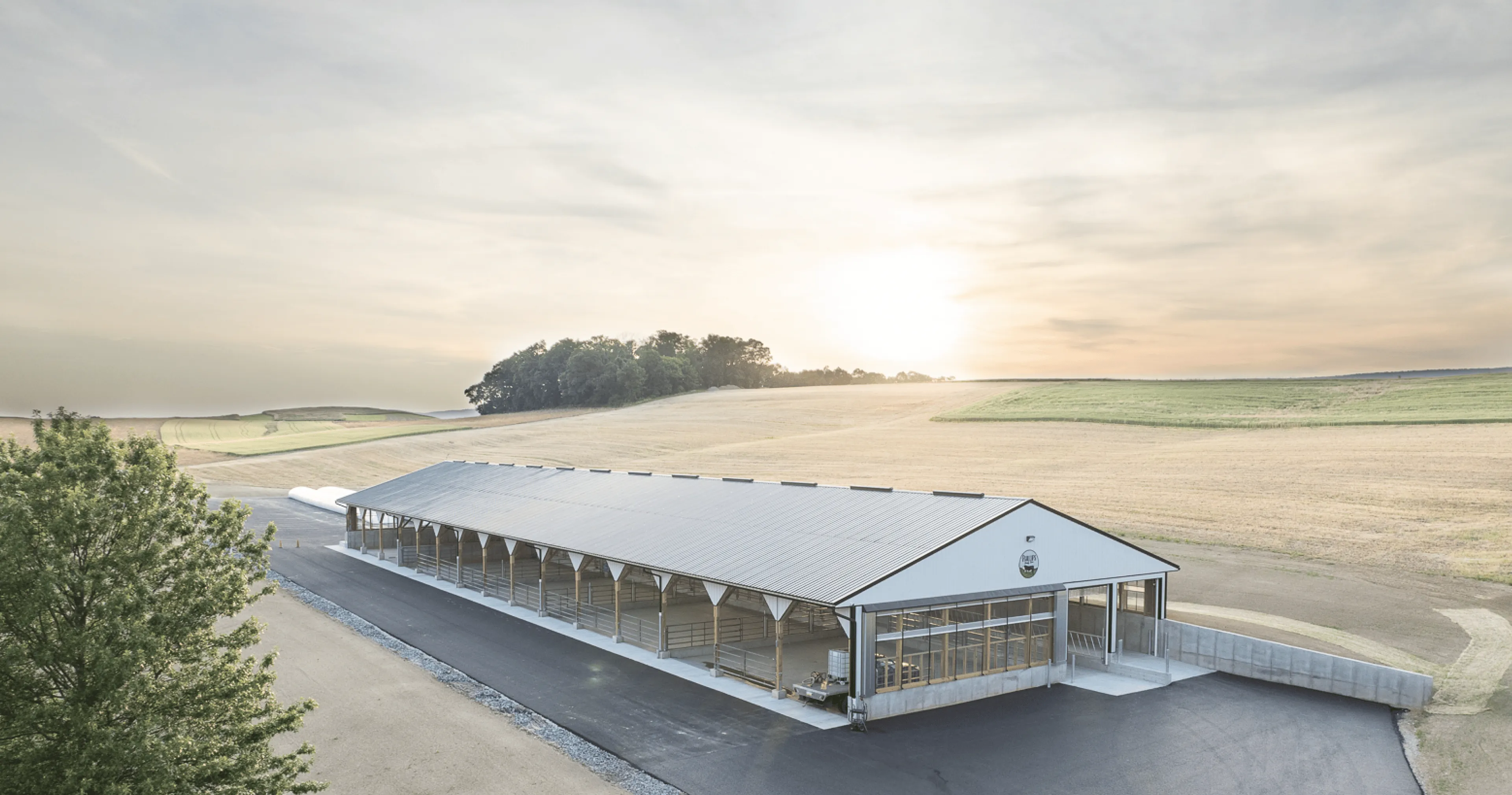 A modern, open-sided agricultural building sits beside a paved road in a rural landscape with rolling fields under a hazy sunrise sky.