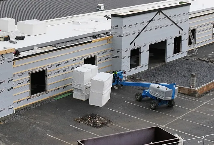 A construction site shows a building under renovation with insulation material stacked outside and a blue boom lift positioned near the entrance.