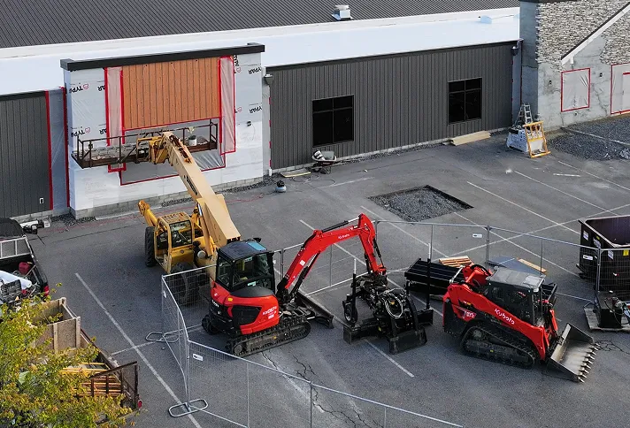 Aerial view of a construction site with three pieces of heavy machinery, fencing, and workers installing a large panel on a building exterior.