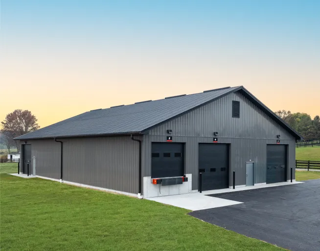 A large gray metal building with three black garage doors and a paved driveway sits on a grassy lot.