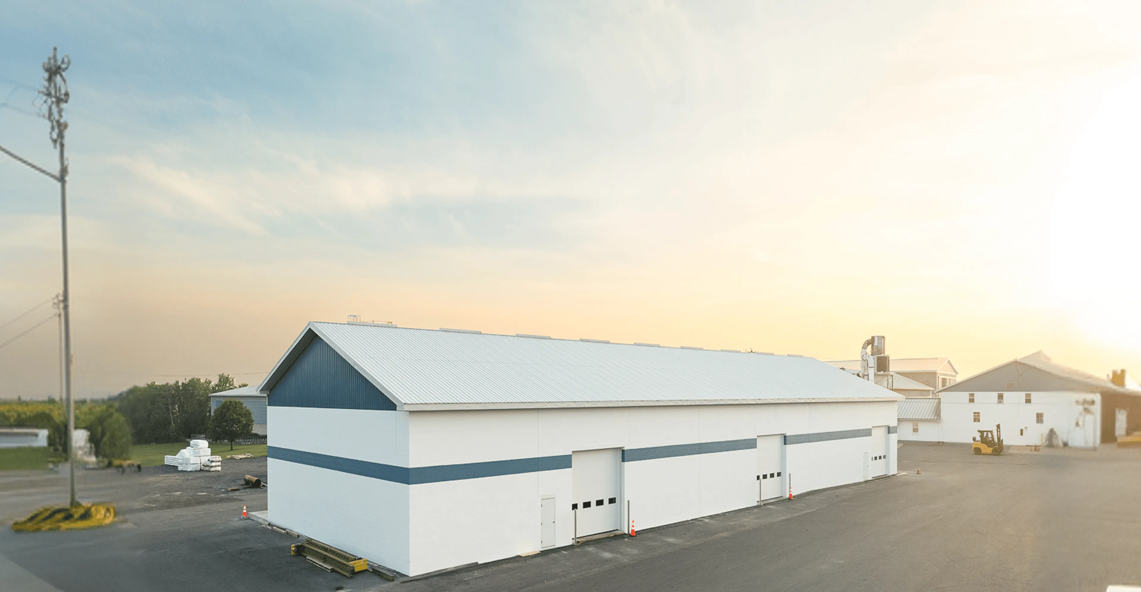 A large white industrial warehouse with blue trim sits on a paved lot under a clear sky, with other buildings and greenery in the background.