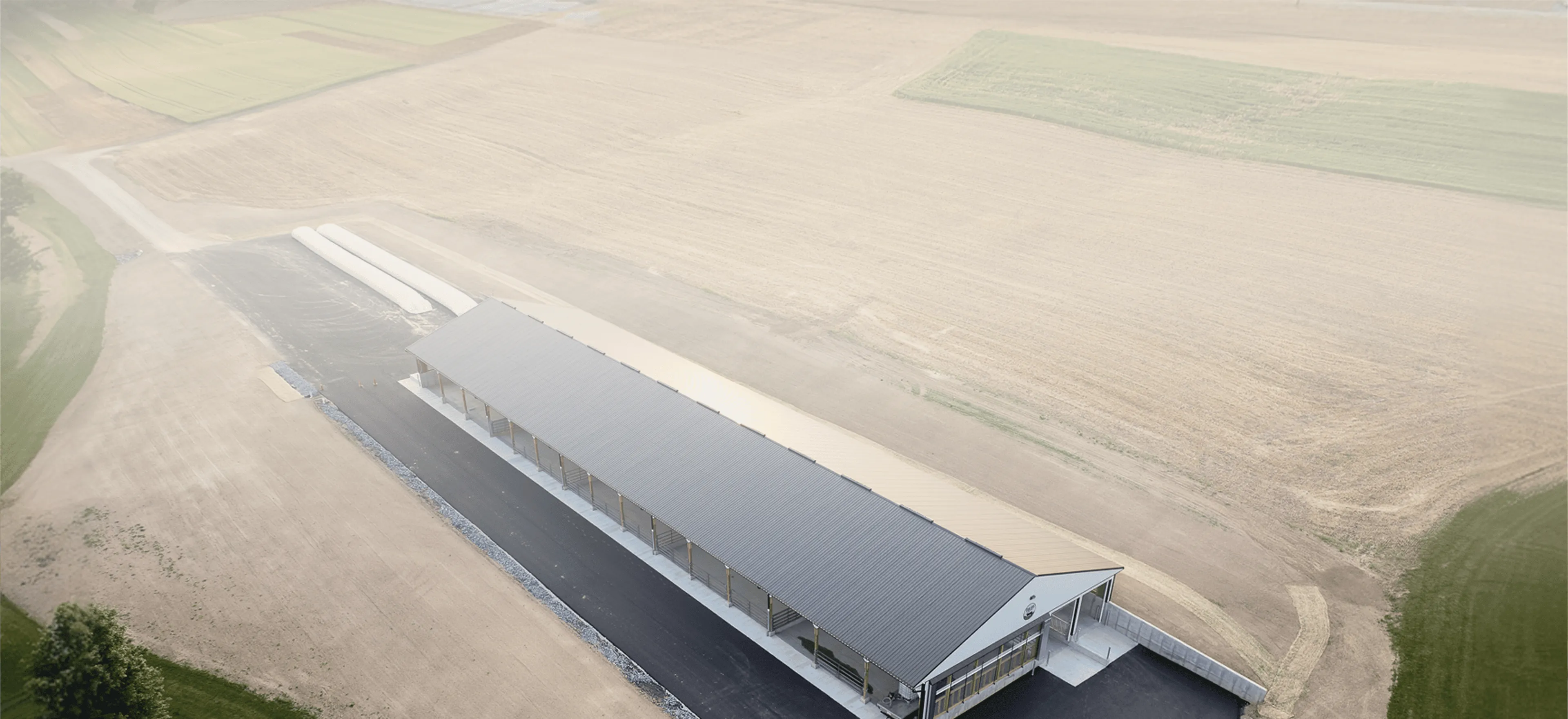 A long, rectangular industrial building with a dark roof sits in the middle of large, tilled farmland.