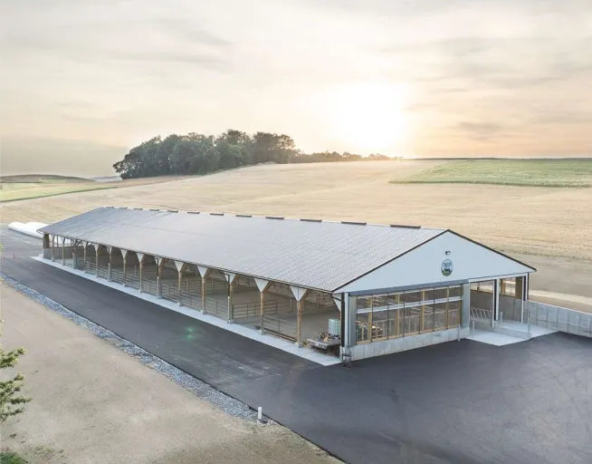A large, modern barn structure sits on a paved area surrounded by open fields.