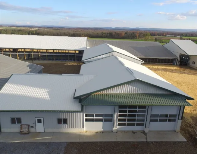 A large agricultural building with green roofs and multiple garage doors, situated in a rural area with open fields and distant hills.