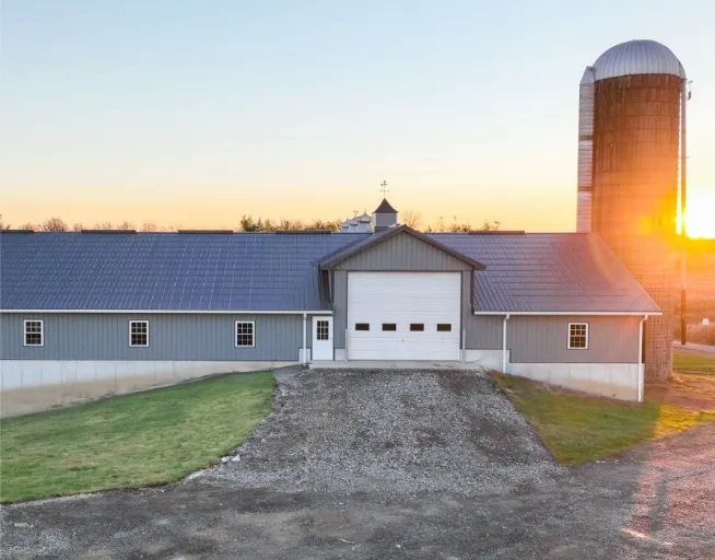 A gray barn with white trim and a large garage door stands next to a tall silo at sunset on a rural property.