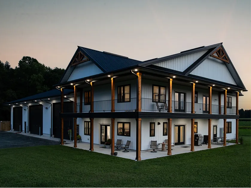 Two-story modern barndominium with a metal roof, large garage bays, upper balcony, and covered ground-level patio, set on a grassy lawn at dusk.