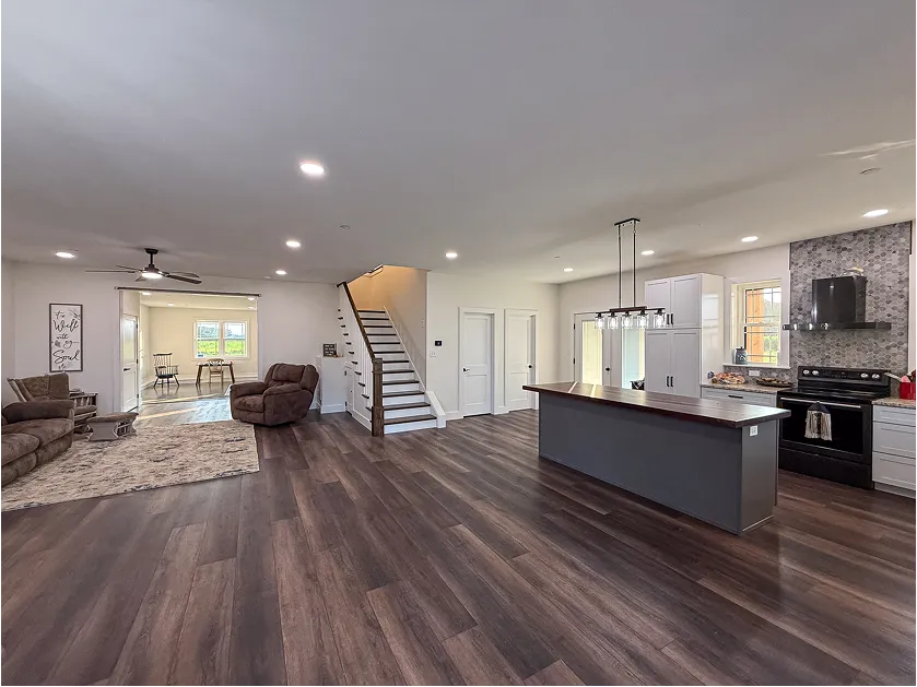 Open-concept living area with dark wood flooring, a kitchen island, modern lighting, stairs, and seating area with recliners and a ceiling fan.