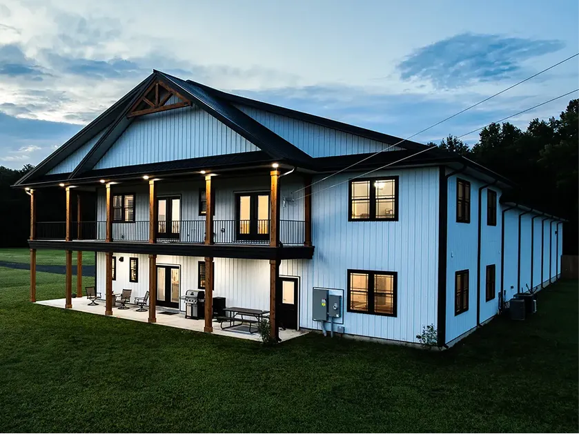 A large two-story modern barn-style house with white siding, black trim, a covered balcony, and outdoor seating, set on a grassy lawn.
