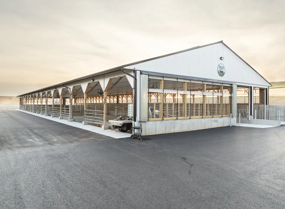 A large, modern, open-sided barn with metal siding and a pitched roof stands next to an asphalt surface.