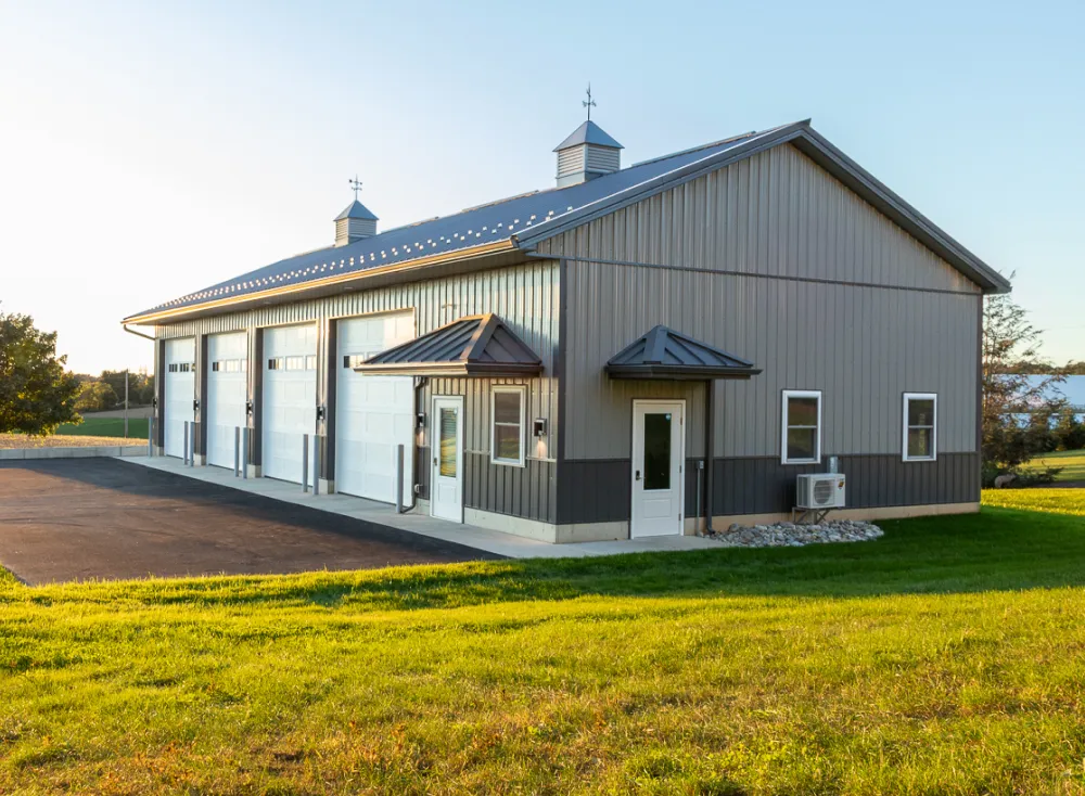 A large metal building with four garage doors and a small office entrance, situated on a paved area and surrounded by grass on a clear day.