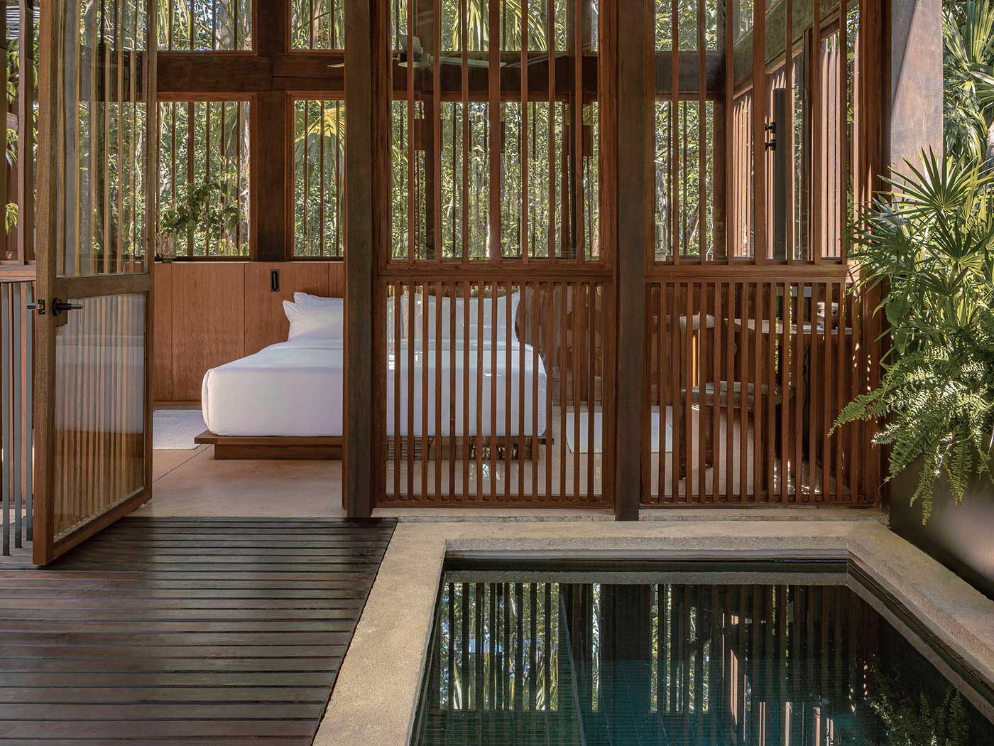 An elegant guest room at La Valise Mazunte Hotel, with a king-size bed facing a wooden terrace and a lush green garden. The interior blends wood walls and natural stone.