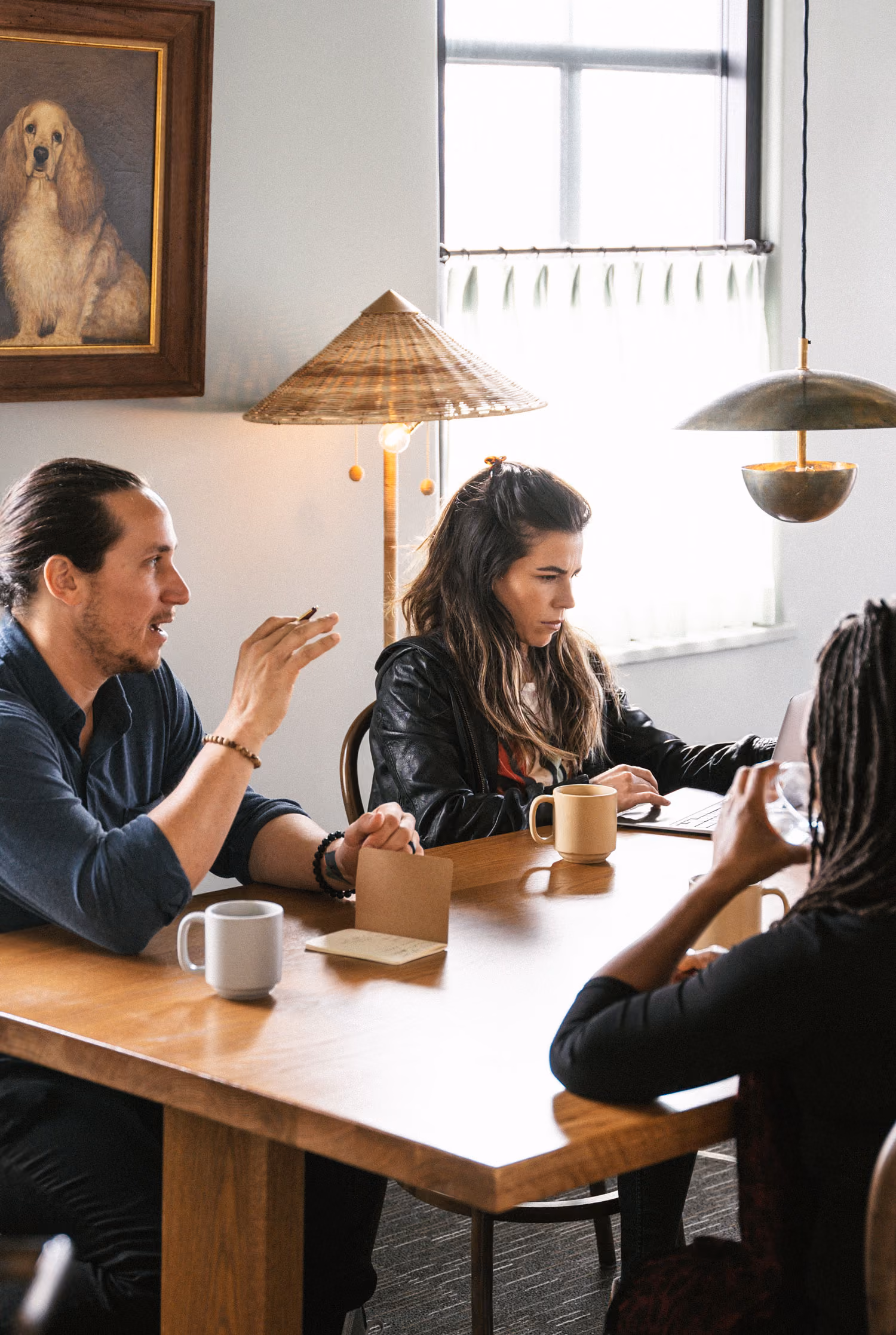 Three people having a meeting around a wooden table with coffee mugs in a bright room.