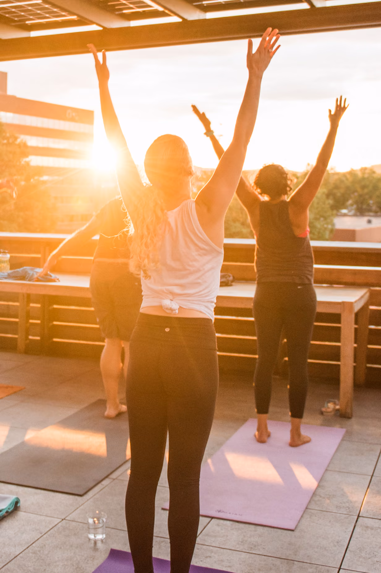 Three people practicing yoga with arms raised on mats outdoors at sunset on a rooftop.