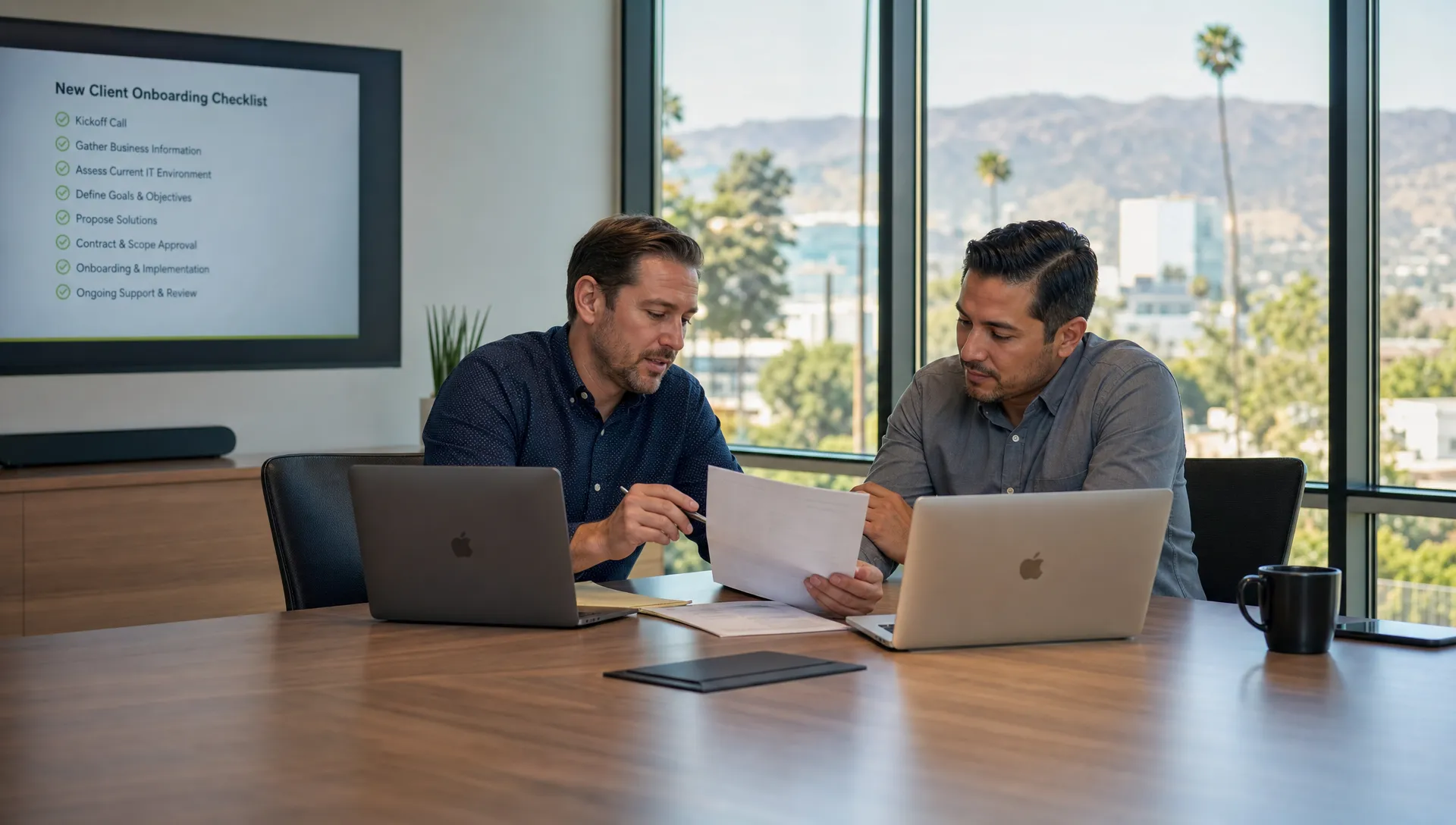 A modern conference room in a Southern California office where a business owner and IT consultant review an onboarding checklist with laptops and printed notes, with the laptop screens facing them, natural daylight, and a clean uncluttered setting.