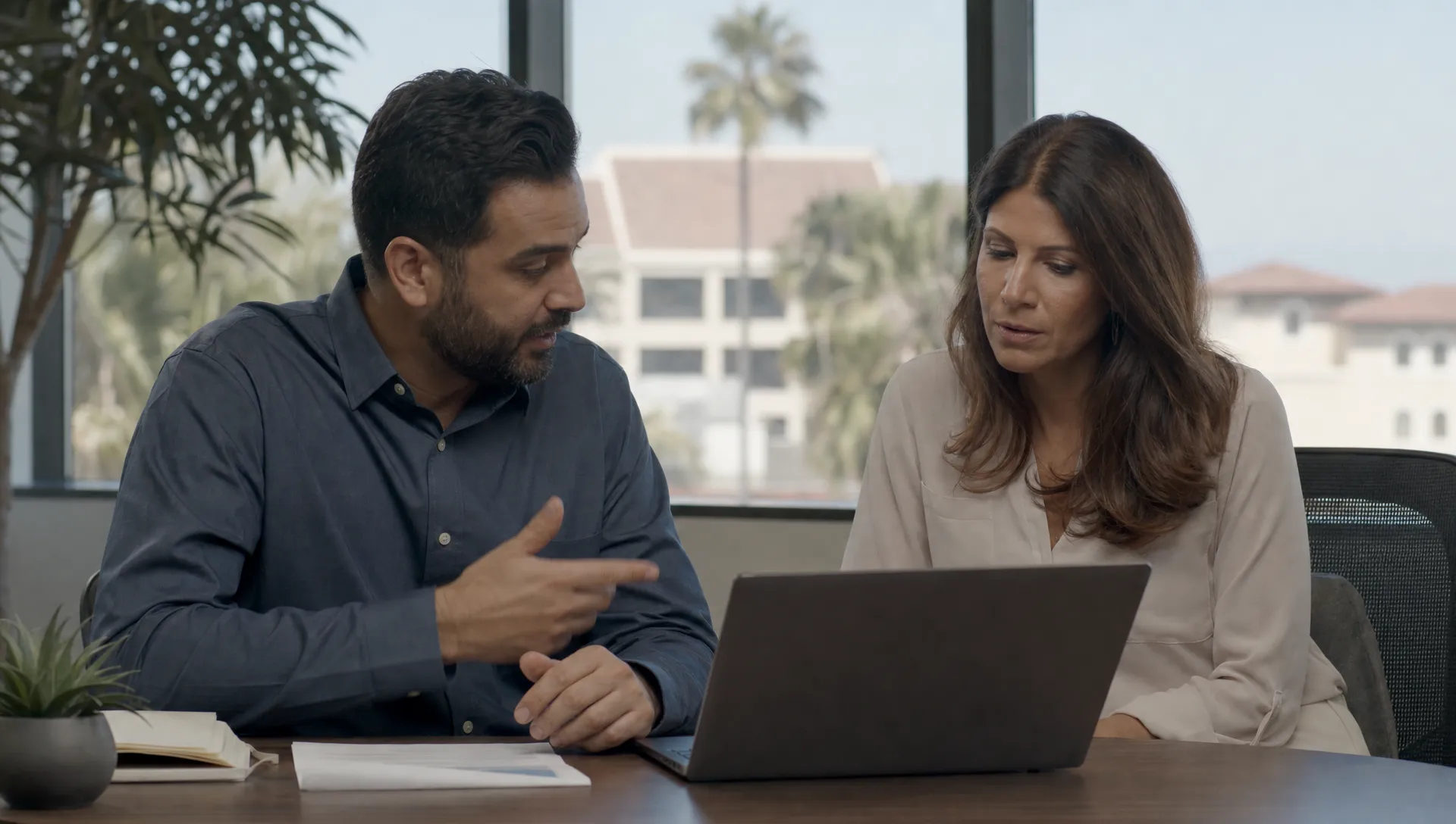 An IT consultant and office manager discuss endpoint protection and recovery planning beside an open laptop in a Southern California office.
