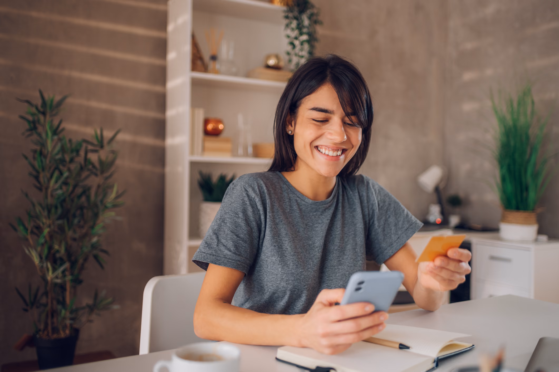 Smiling woman holding a credit card and smartphone, sitting at a desk with a notebook and coffee.