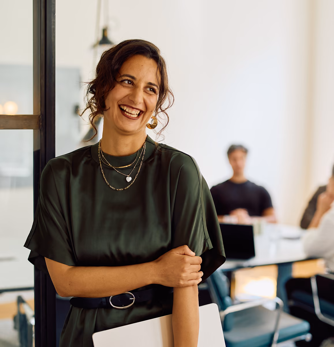Smiling woman standing in an office with a laptop, with coworkers in the background.