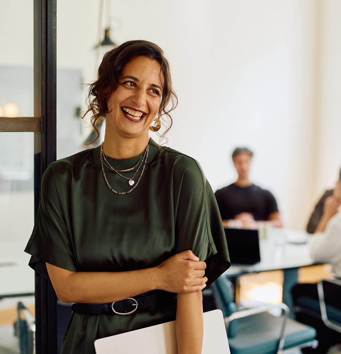 Smiling woman standing in an office with a laptop, with coworkers in the background.