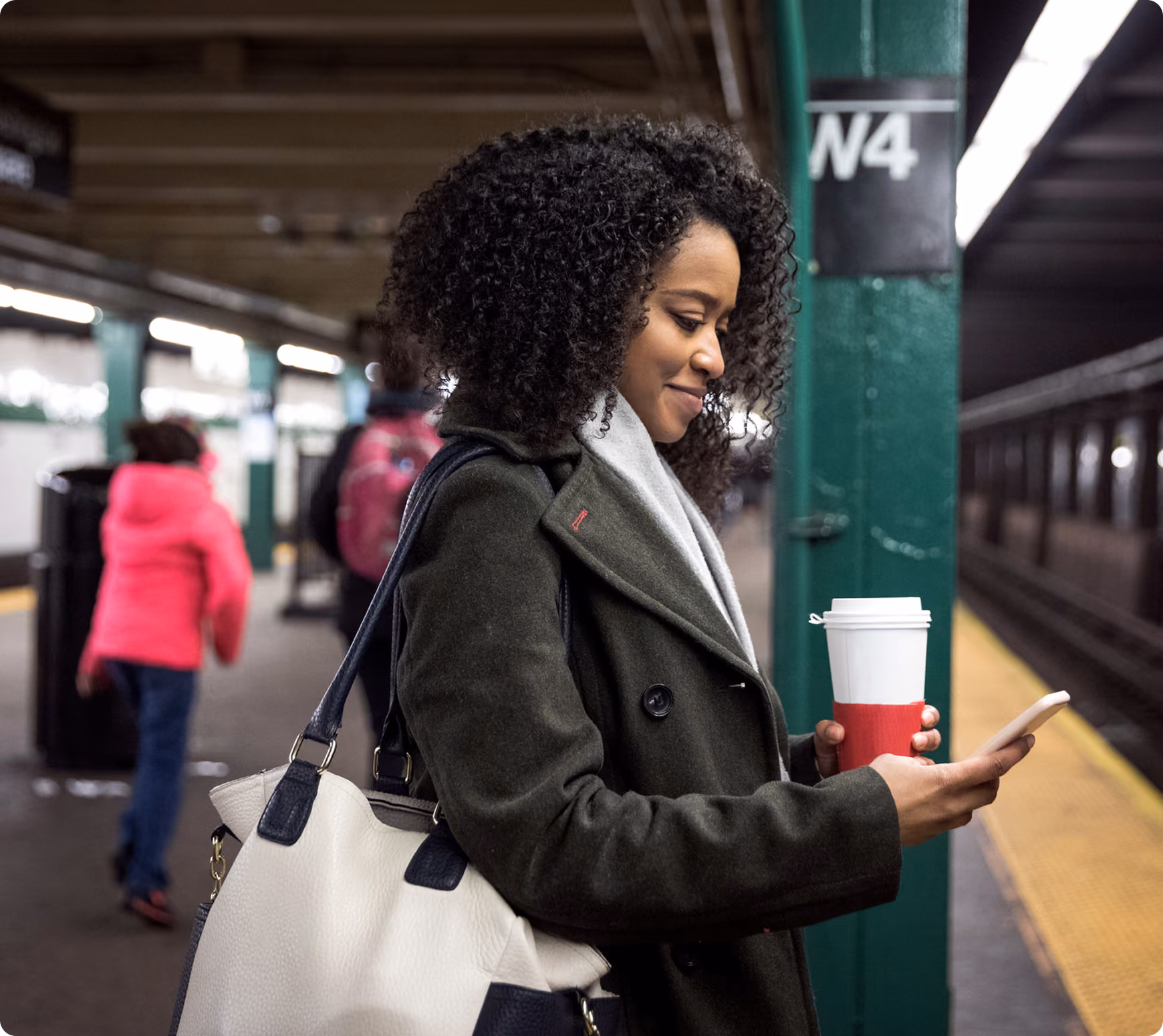 Woman with curly hair checks phone and holds coffee cup on a subway platform near a green pillar marked W4.
