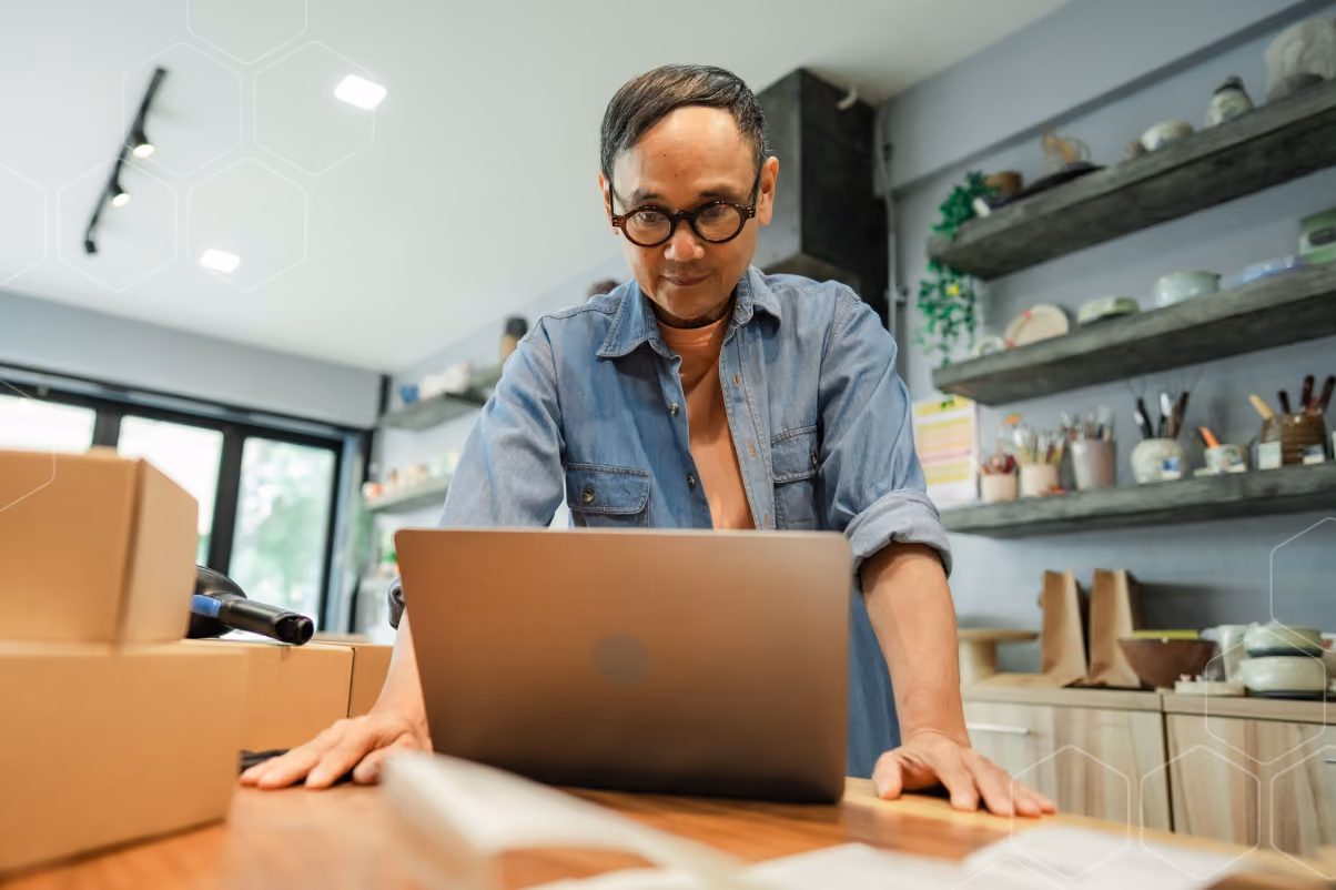 Man wearing glasses working on a laptop in a small business workspace with shelves and boxes around.