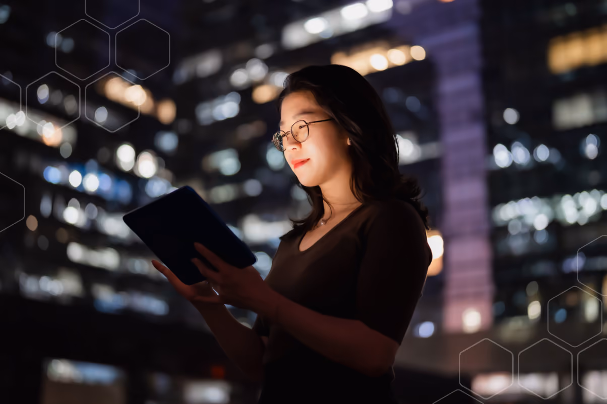 Woman wearing glasses using a tablet with city lights blurred in the background at night.