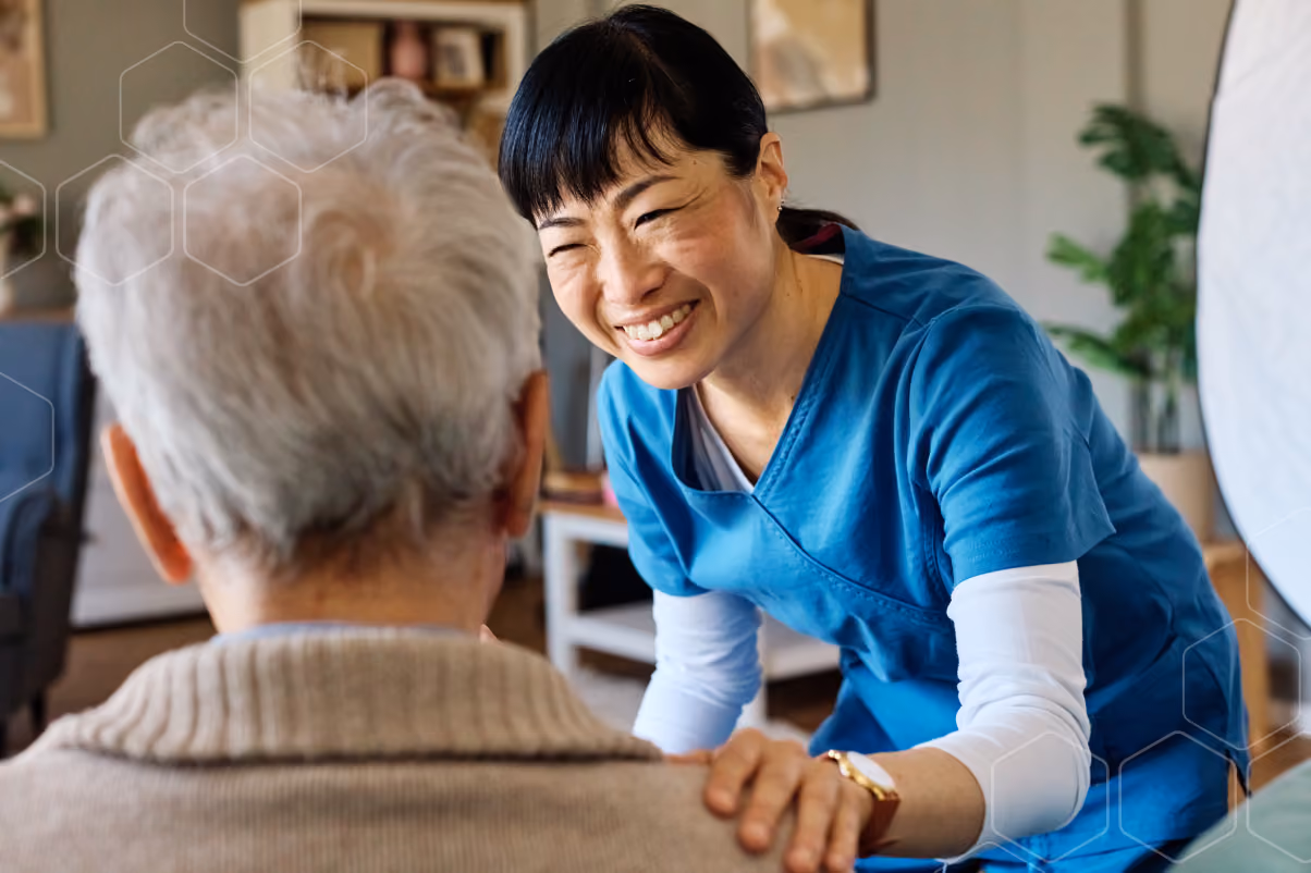 Smiling female healthcare worker in blue scrubs comforts elderly person with white hair.