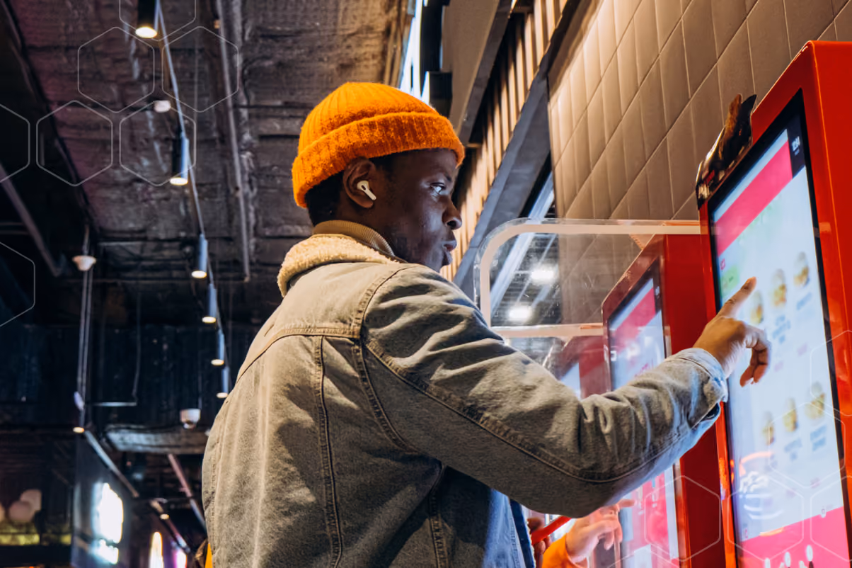 Person in orange beanie and denim jacket uses a touchscreen kiosk in a modern indoor space.
