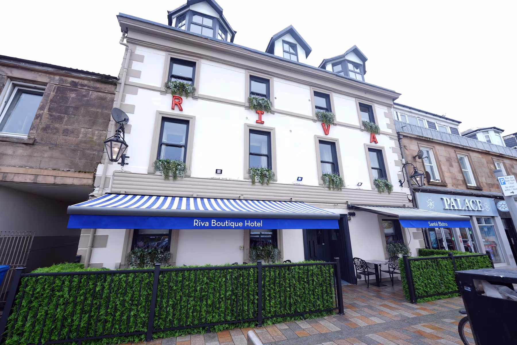 Exterior of Riva Boutique Hotel in Helensburgh, showing the white Victorian facade with red neon RIVA lettering, blue striped awning, and Santé Bistro entrance below.