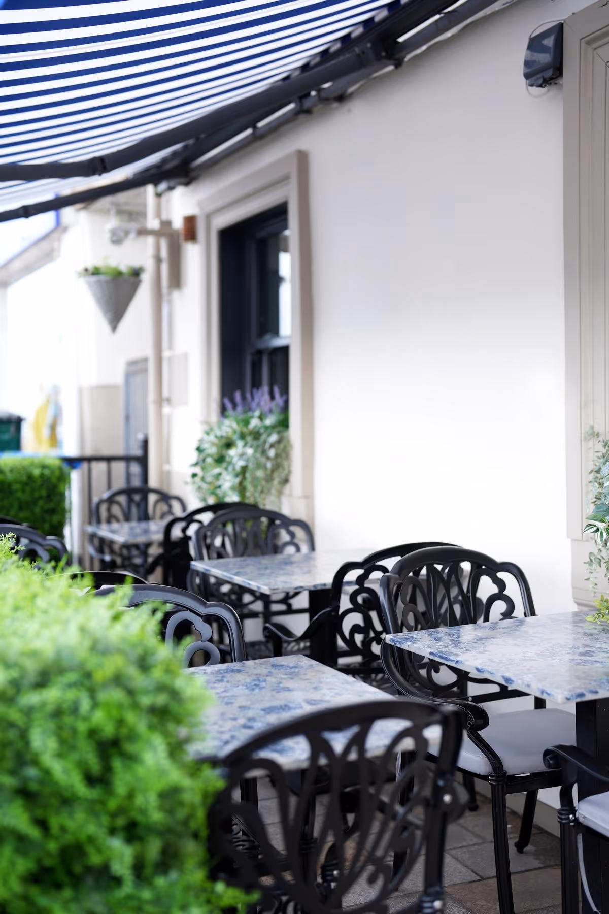 Close-up of the Riva Helensburgh outdoor terrace with marble-topped tables and ornate black chairs beneath a blue and white striped canopy, with green hedging in the foreground.