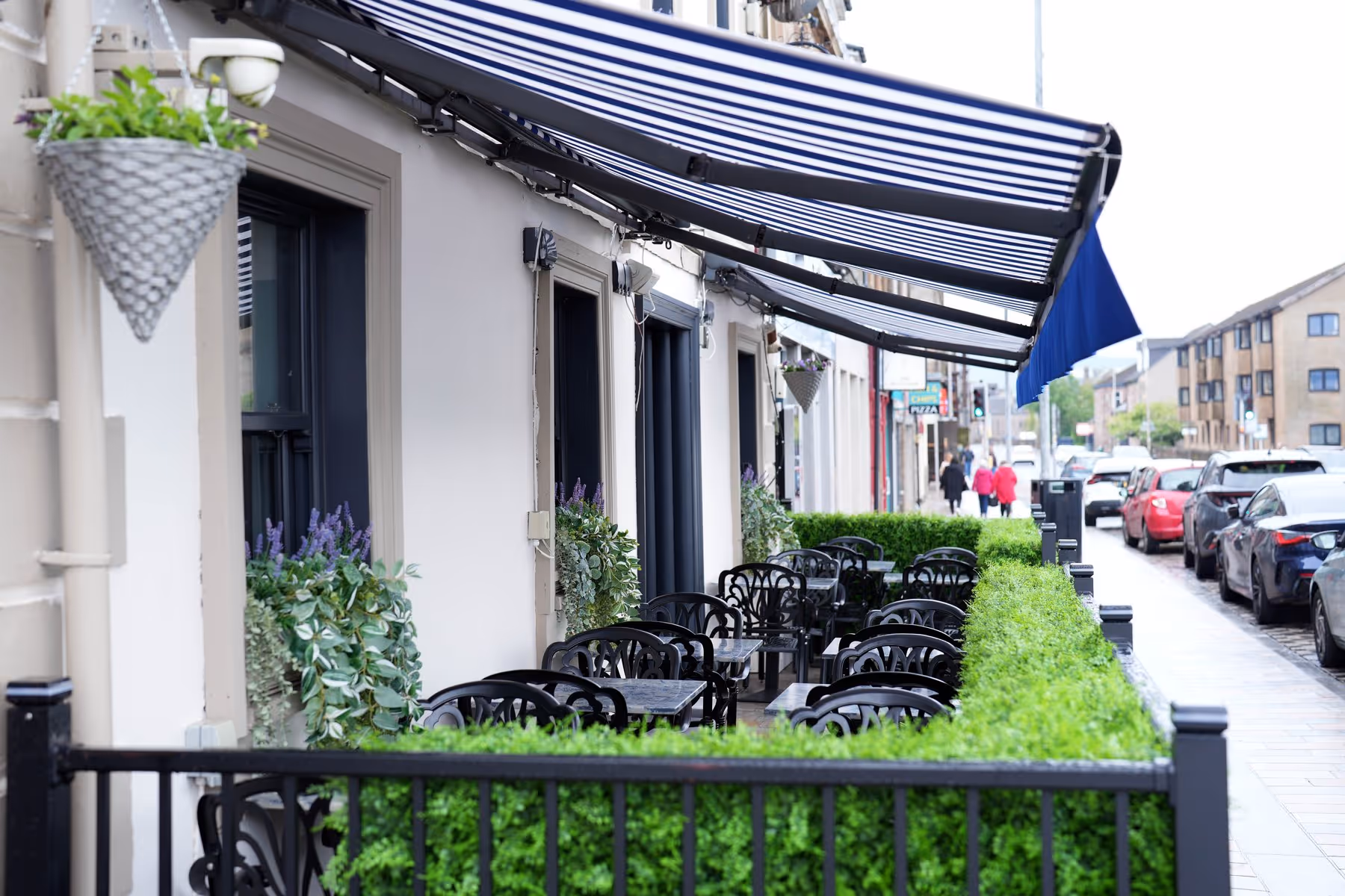 Outdoor seating area at Riva Helensburgh with black cast-iron tables and chairs beneath a blue and white striped awning, lined with trimmed hedges along the pavement.