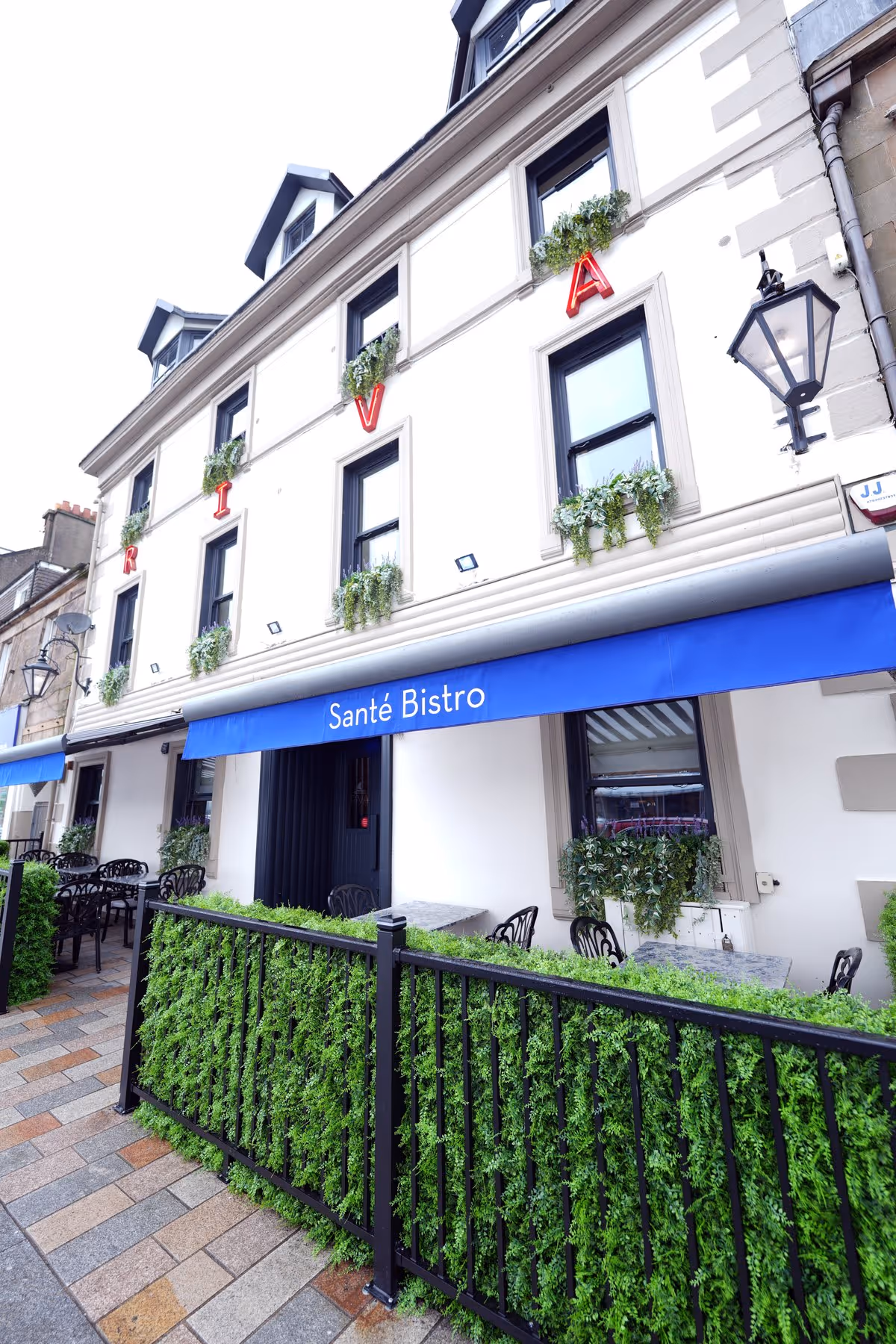 Front facade of Riva Helensburgh showing the Santé Bistro entrance with a blue awning, hanging basket plants, and the neon RIVA letters on the upper floors.