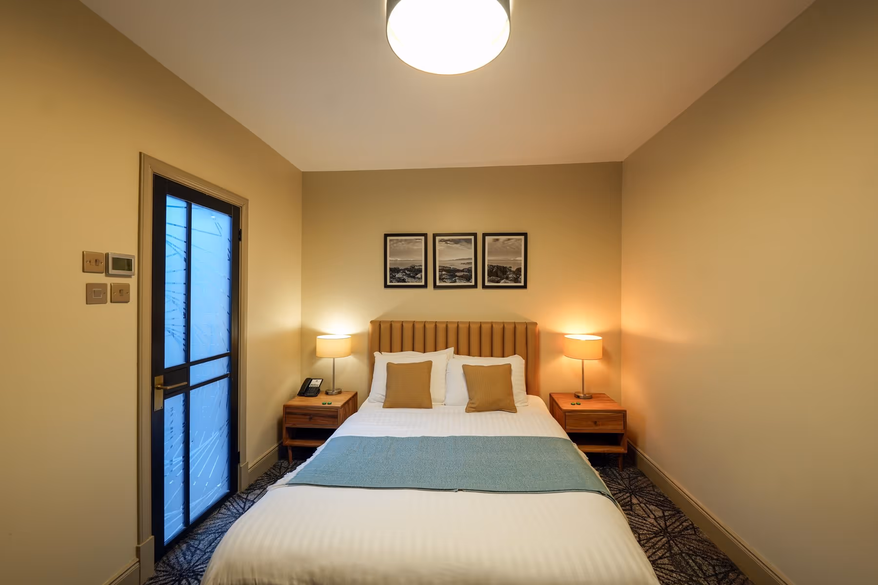 King Room at Riva Helensburgh — full room view showing the kingsize bed with tan headboard, twin bedside lamps, and a frosted glass en-suite door.